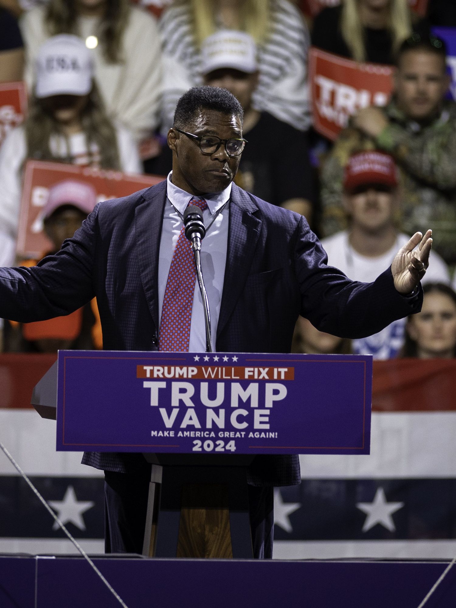 Herschel Walker, a Black man with glasses and a black suit, speaks behind a blue lectern at a Trump rally in Georgia.