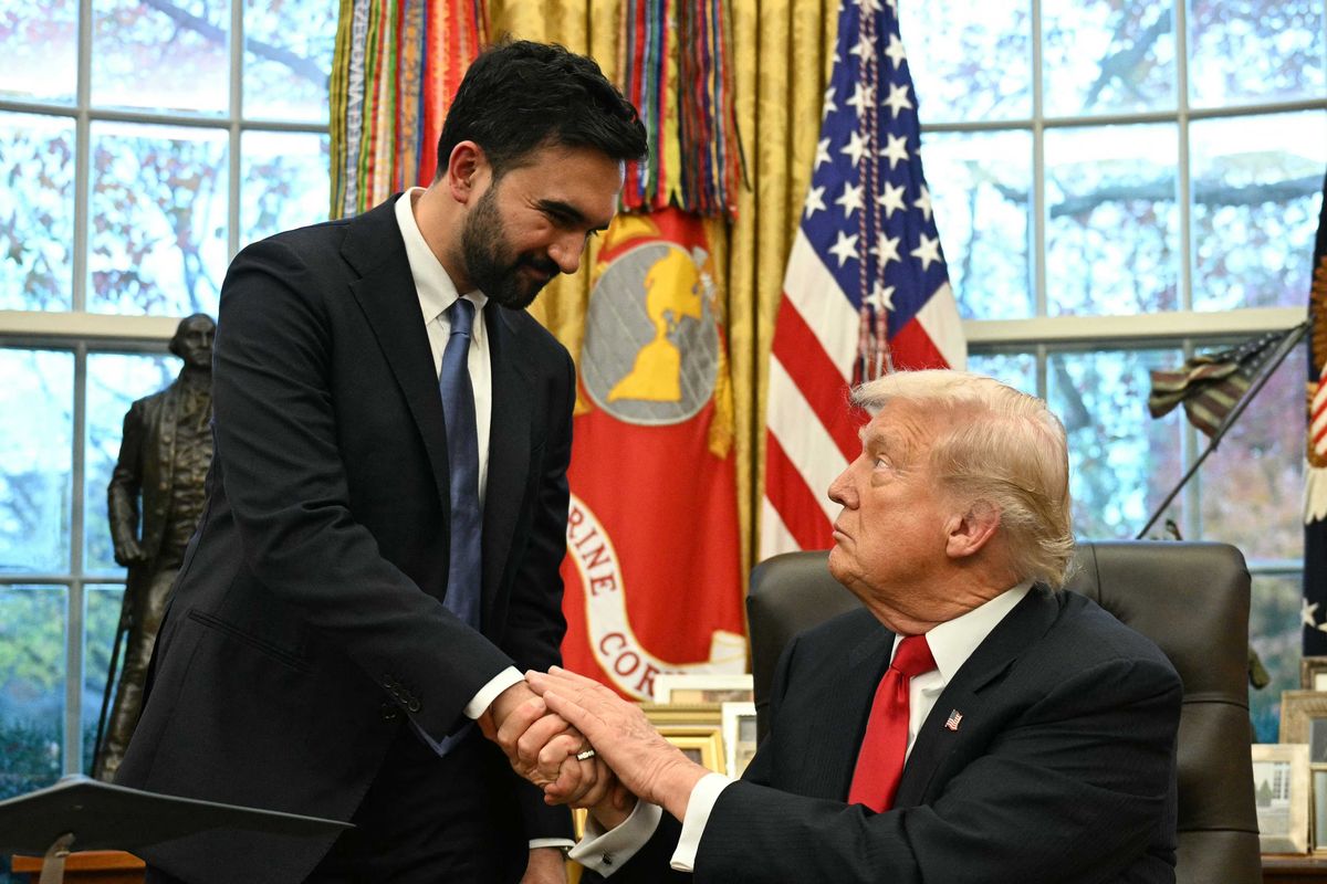 In the Oval Office, Zohran Mamdani, standing left, shakes the hand of Donald Trump, sitting down in a black chair behind his desk.