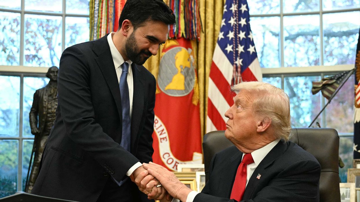 In the Oval Office, Zohran Mamdani, standing left, shakes the hand of Donald Trump, sitting down in a black chair behind his desk.