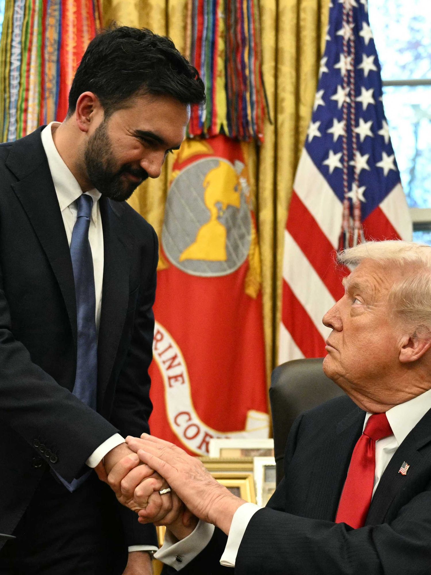 In the Oval Office, Zohran Mamdani, standing left, shakes the hand of Donald Trump, sitting down in a black chair behind his desk.