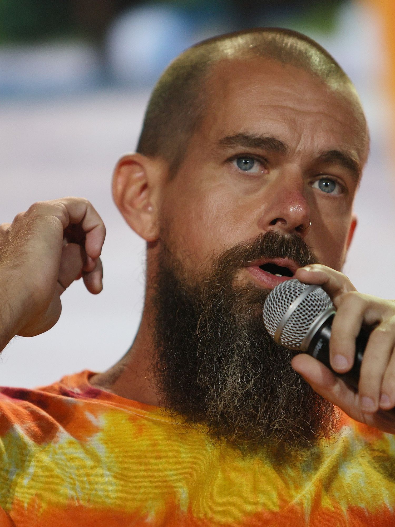 Jack Dorsey, a bearded white man with a shaved head and yellow tie-dye t-shirt, speaks into a microphone in his left hand.