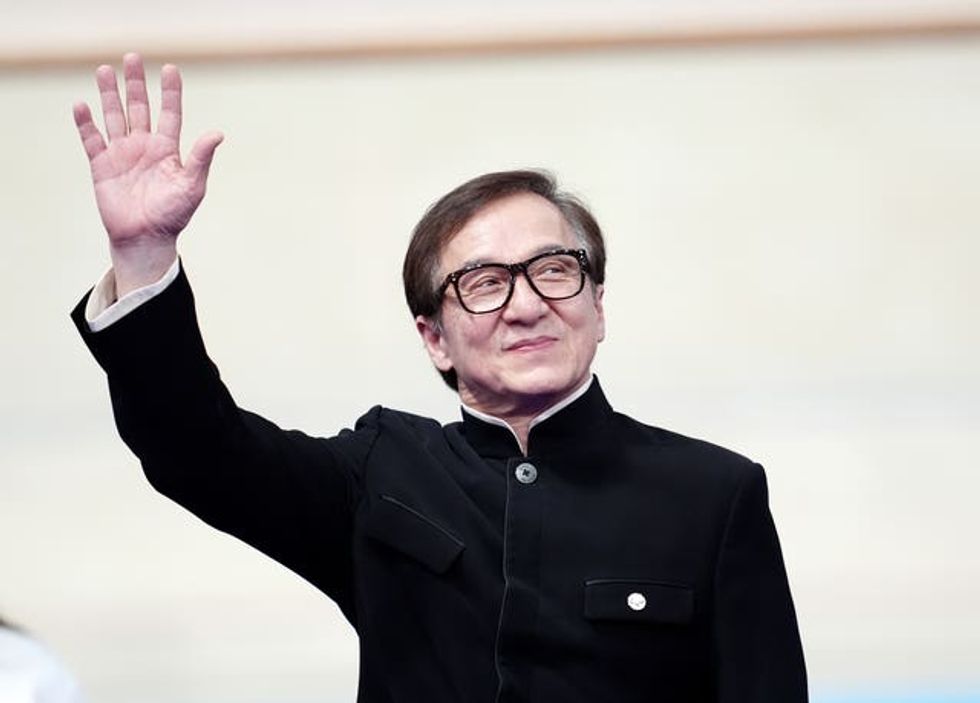 Jackie Chan, in a black jacket, waves