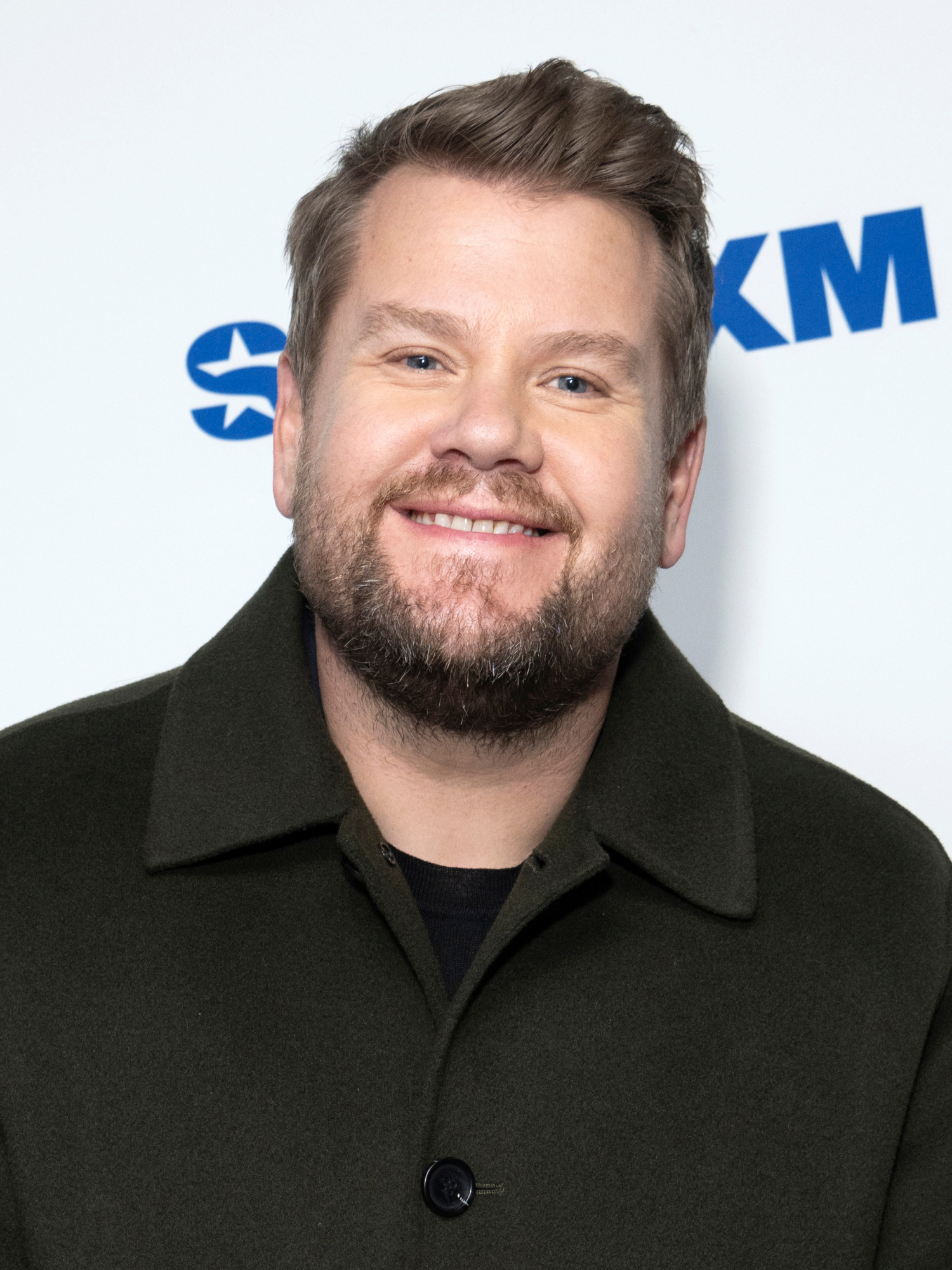 James Corden, a white man with short brown hair and a black coat, smiles in front of a white posterboard.
