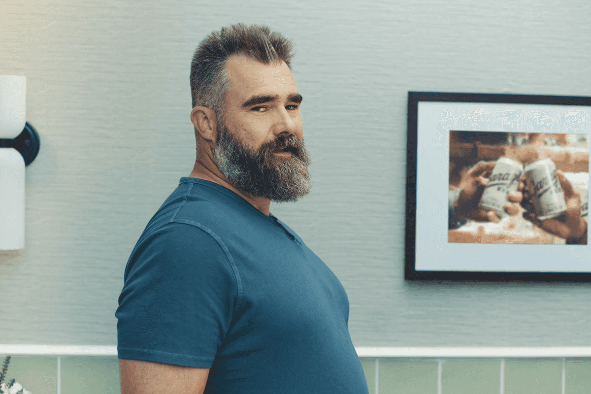 Jason Kelce, a bearded white man with short black hair and a blue T-shirt, looks side on at the camera inside a bathroom.