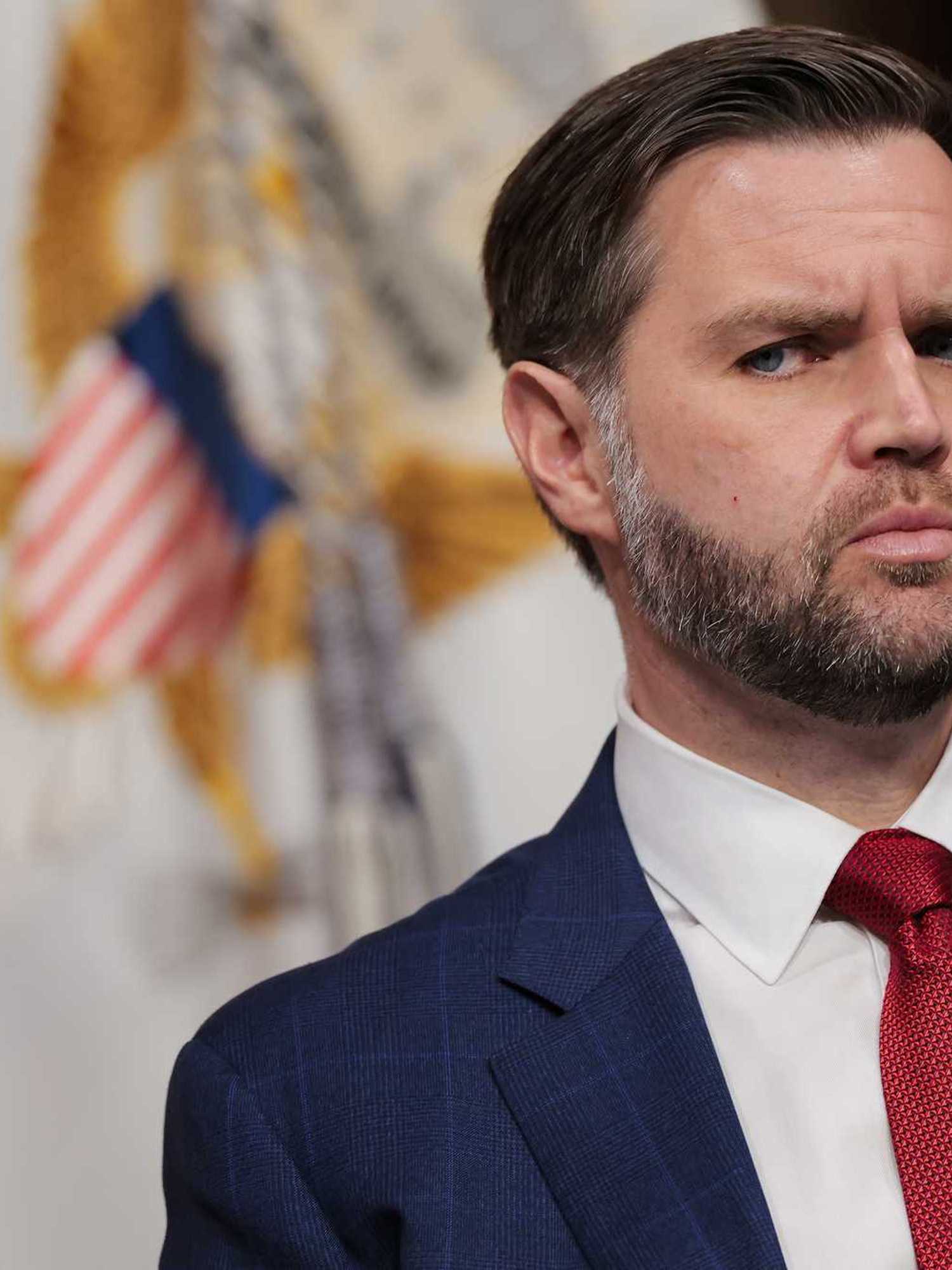 JD Vance in a blue suit and red tie, looking to his right with several flags behind him.
