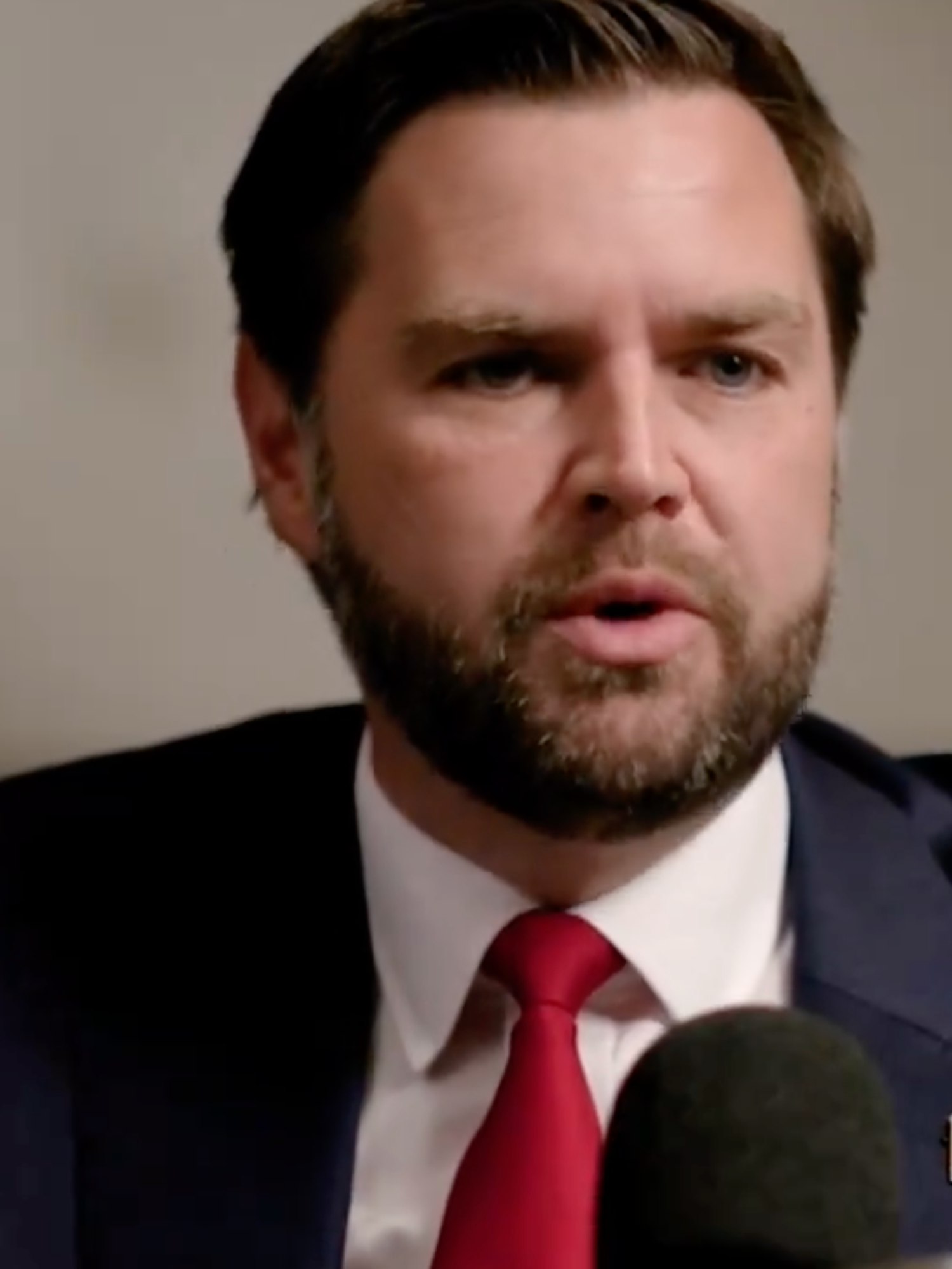 JD Vance in a navy suit speaking into a microphone, and talking to a journalist off-camera.