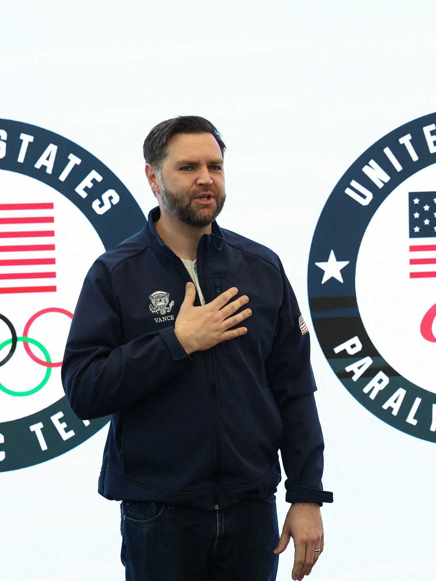 JD Vance places hand on chest as he stands in front of signage for the United States Olympic and Paralympic teams.