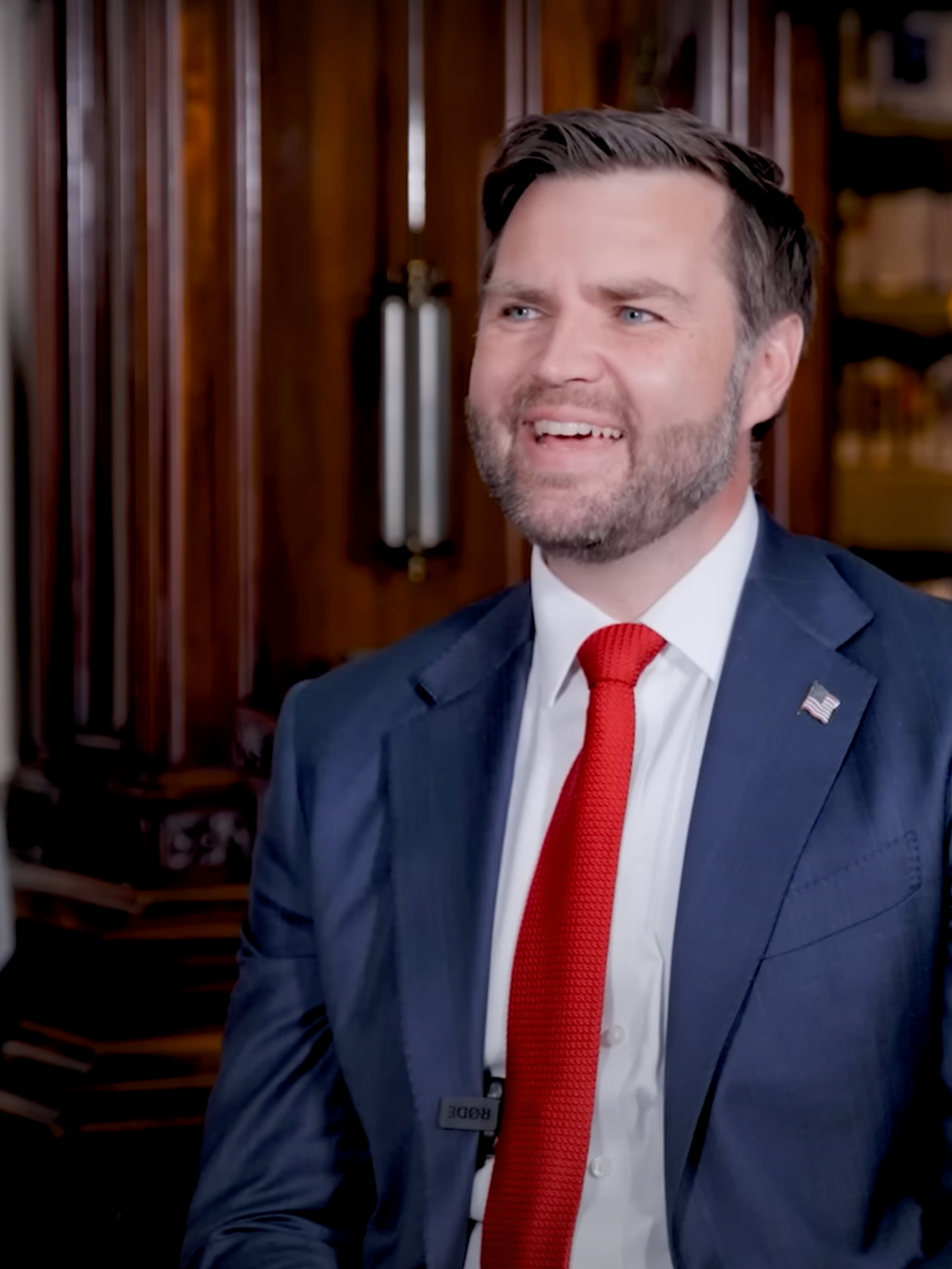 JD Vance smiling, sat in front of two American flags. He's on a podcast.