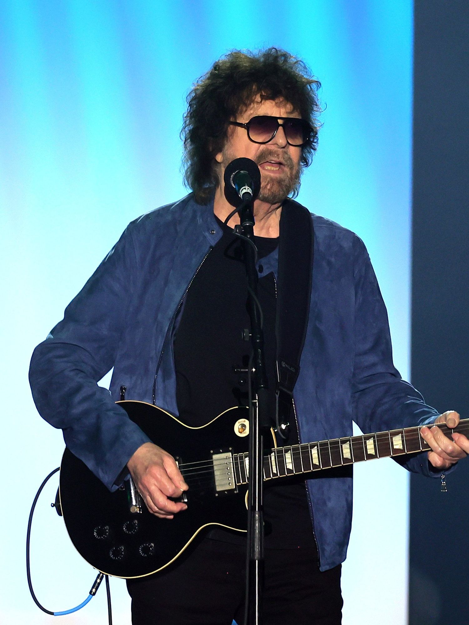 Jeff Lynne, a bearded white man with sunglasses, black curly hair and a blue jacket, plays an electric guitar on a stage.