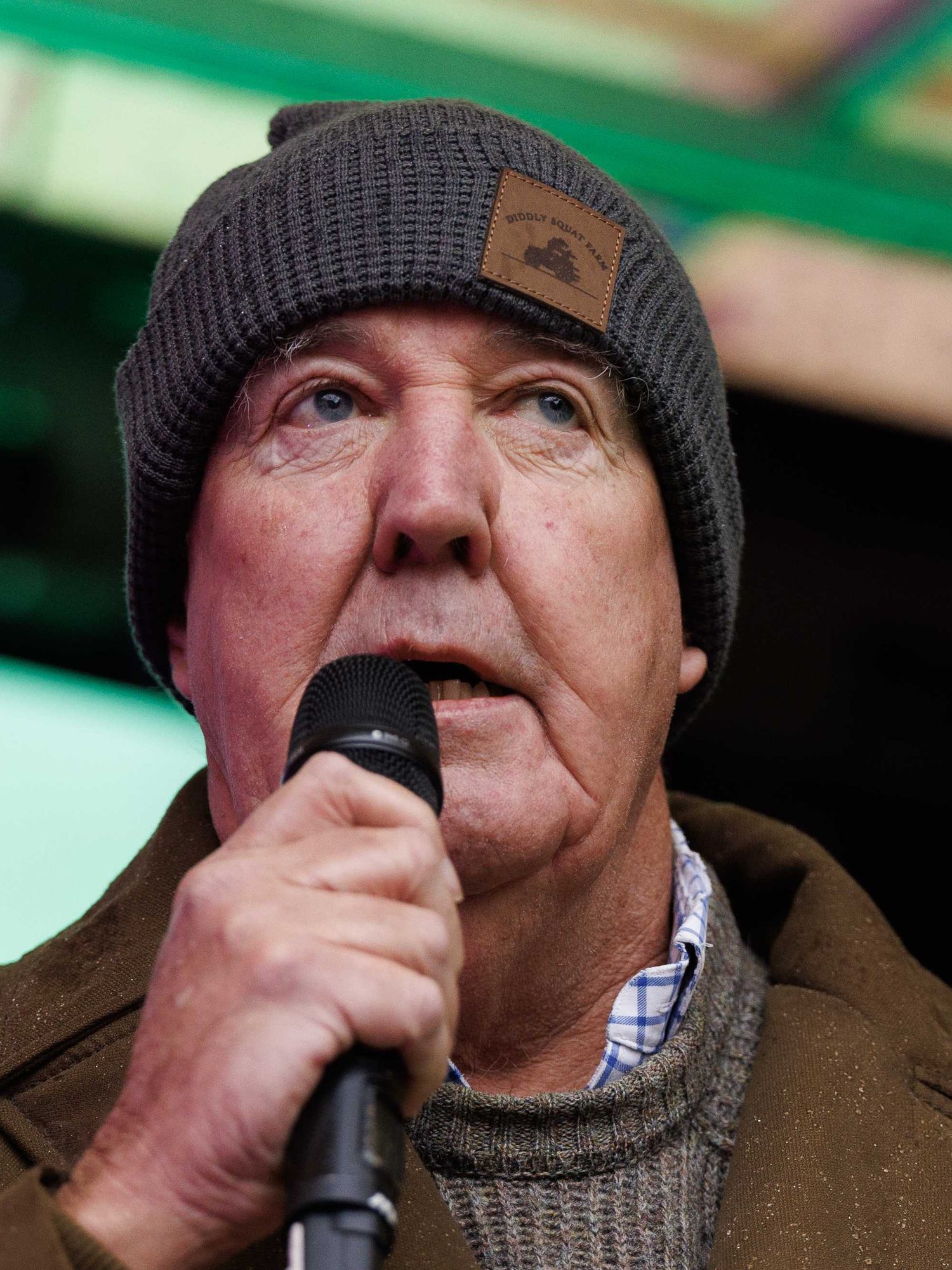 Jeremy Clarkson, in a grey beanie and brown coat, speaks into a microphone.