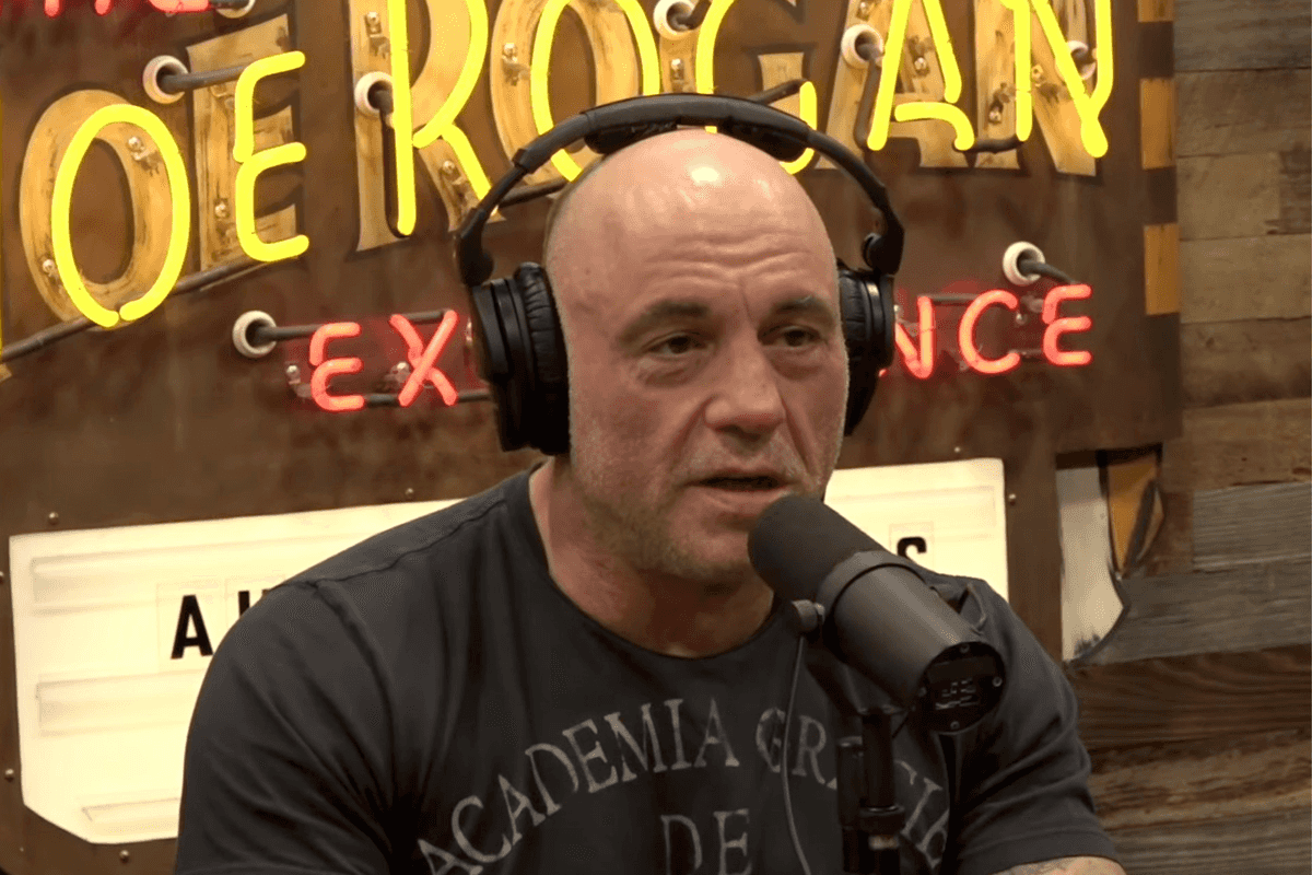Joe Rogan, a bald white man in a grey shirt, wearing headphones and speaking into a microphone as he hosts The Joe Rogan Experience podcast.