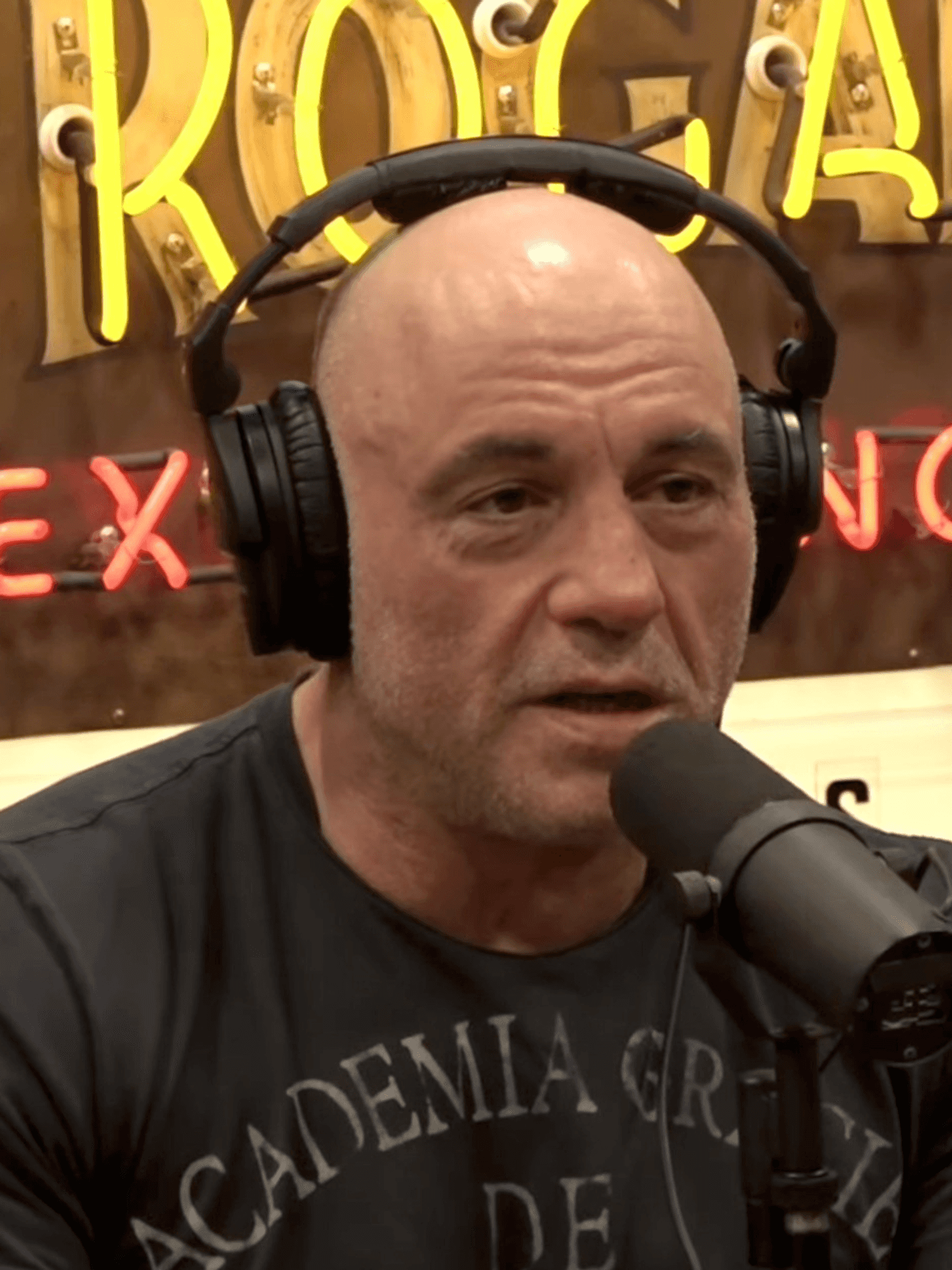 Joe Rogan, a bald white man in a grey shirt, wearing headphones and speaking into a microphone as he hosts The Joe Rogan Experience podcast.
