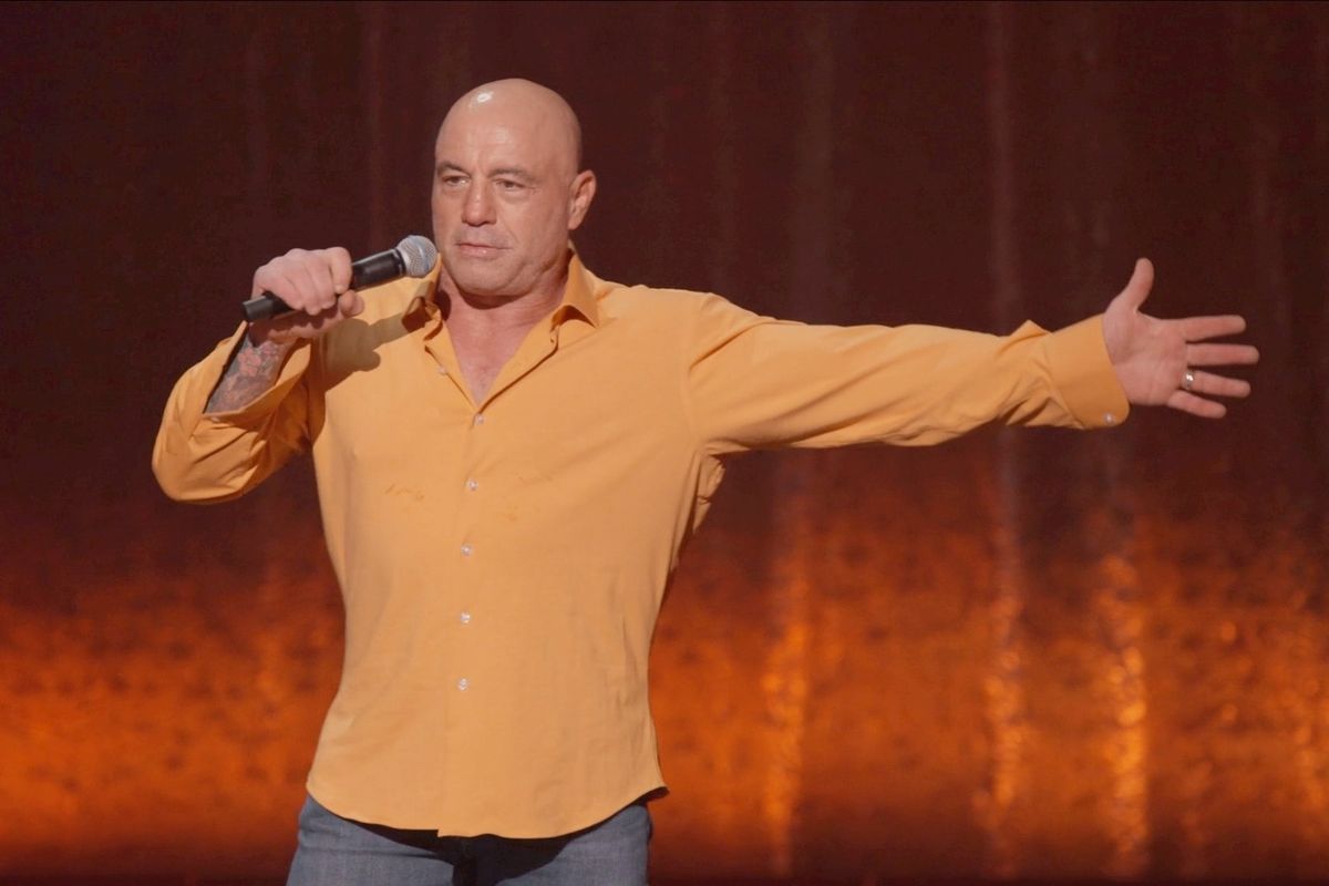Joe Rogan wears a yellow shirt and holds a microphone during his stand-up show