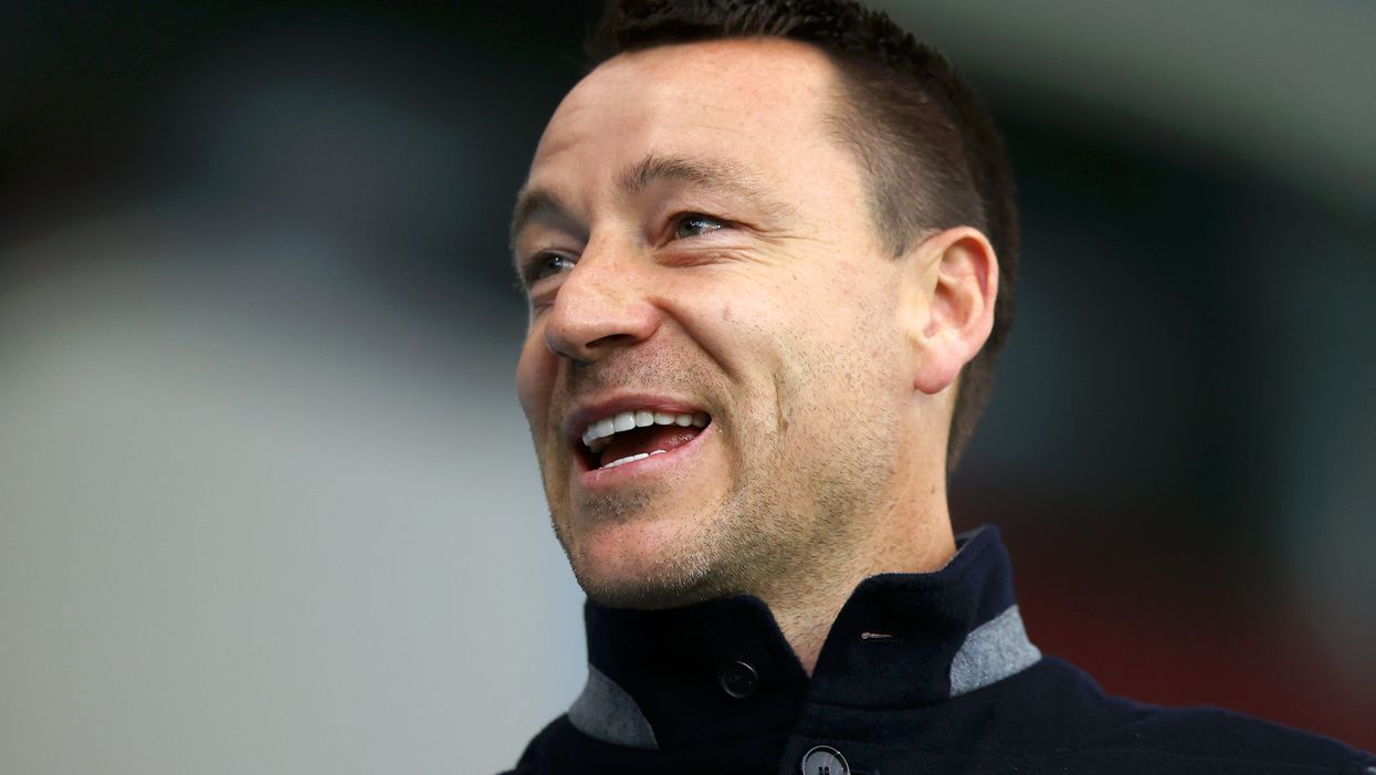 John Terry, a white man with short black hair, smiles as he looks at something to his right. He wears a black collared jumper.