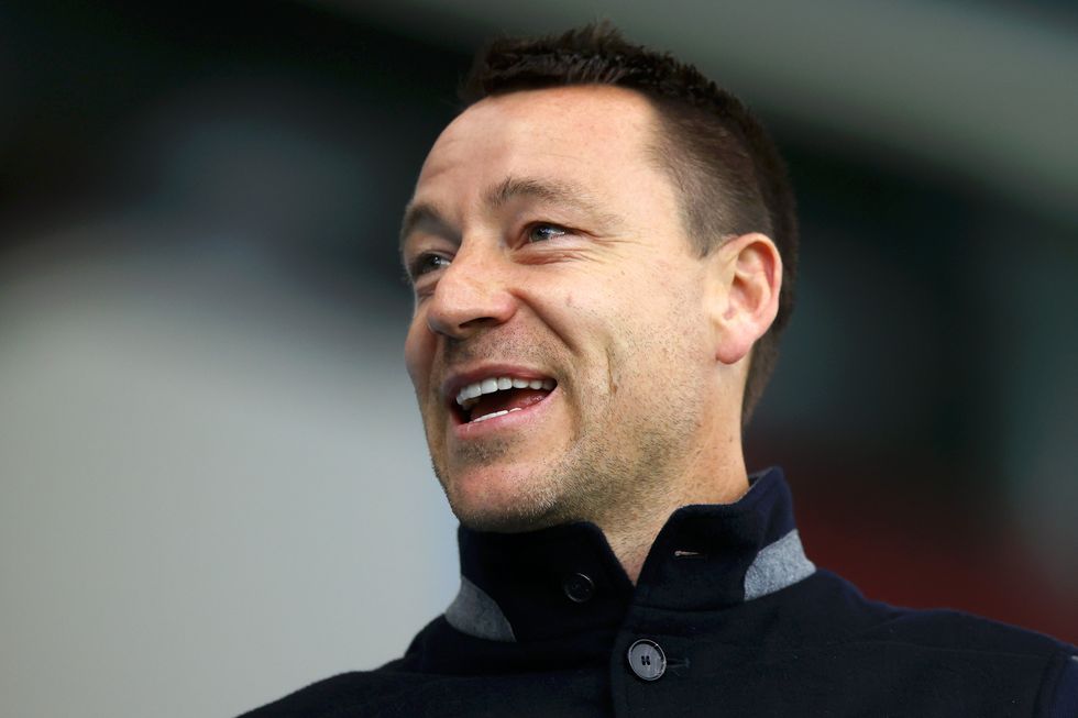 John Terry, a white man with short black hair, smiles as he looks at something to his right. He wears a black collared jumper.
