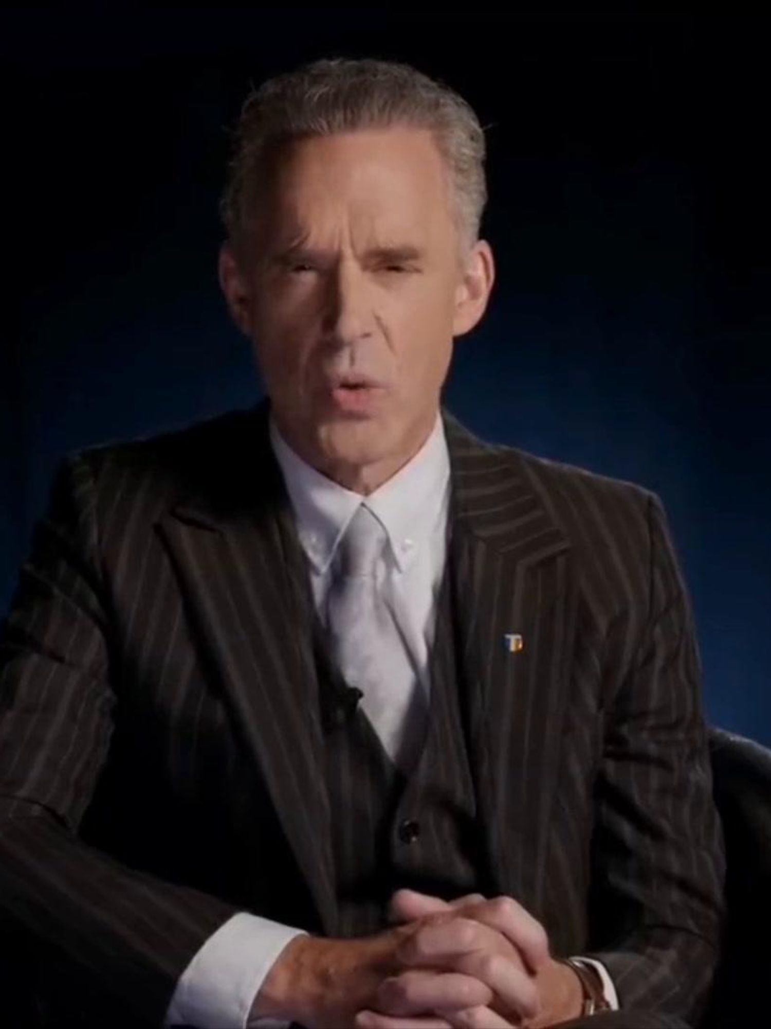 Jordan Peterson, a white man with short grey hair and a black suit, addresses the camera sat down in a black armchair.