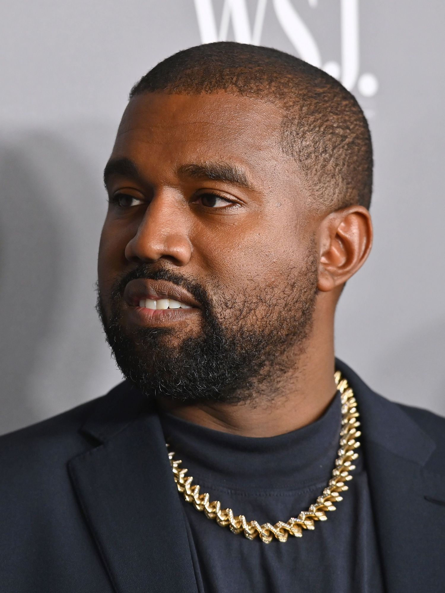 Kanye West in a black suit with a gold neckchain, standing in front of a black posterboard.