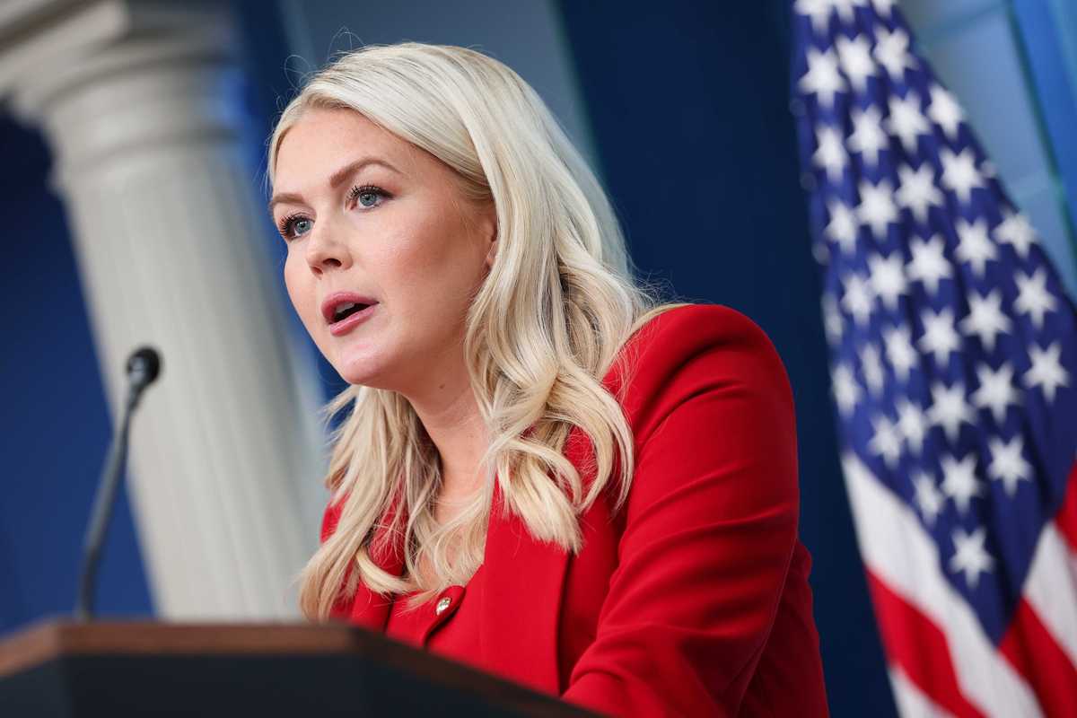 Karoline Leavitt, a white woman with long, wavy blonde hair and a red jacket, speaks to reporters in the White House.