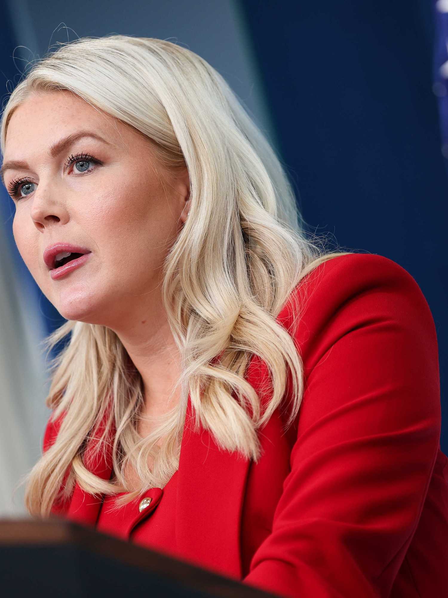 Karoline Leavitt, a white woman with long, wavy blonde hair and a red jacket, speaks to reporters in the White House.