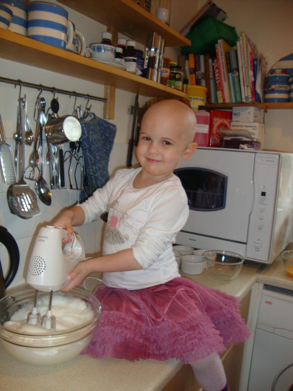 Katie Oates, having lost her hair due to cancer treatment, smiling as a child while using a whisk in a mixing bowl