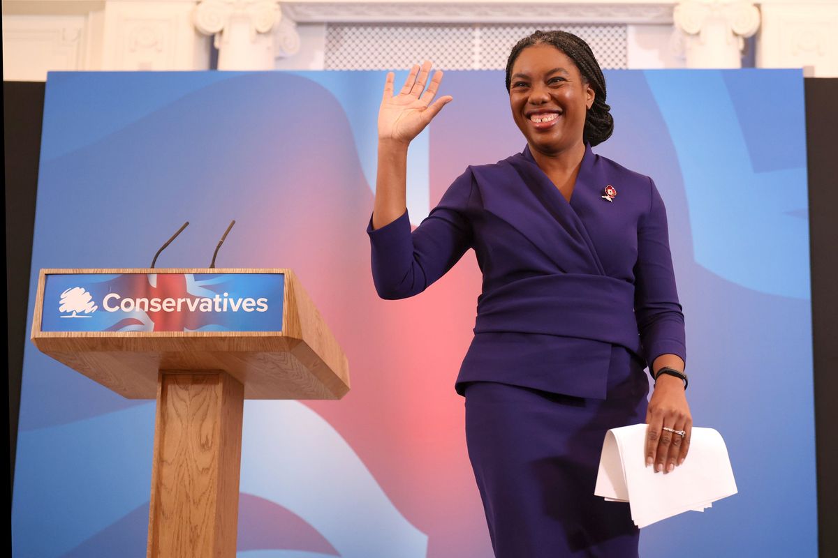 Kemi Badenoch smiles and waves to Tories on a stage with a wooden 'Conservatives' lectern and UK flag behind her.