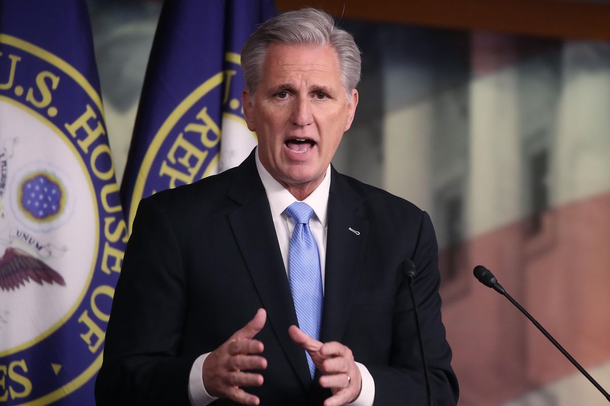 Kevin McCarthy, a white man with short grey hair and a black suit, speaks behind a wooden letter and two blue flags.