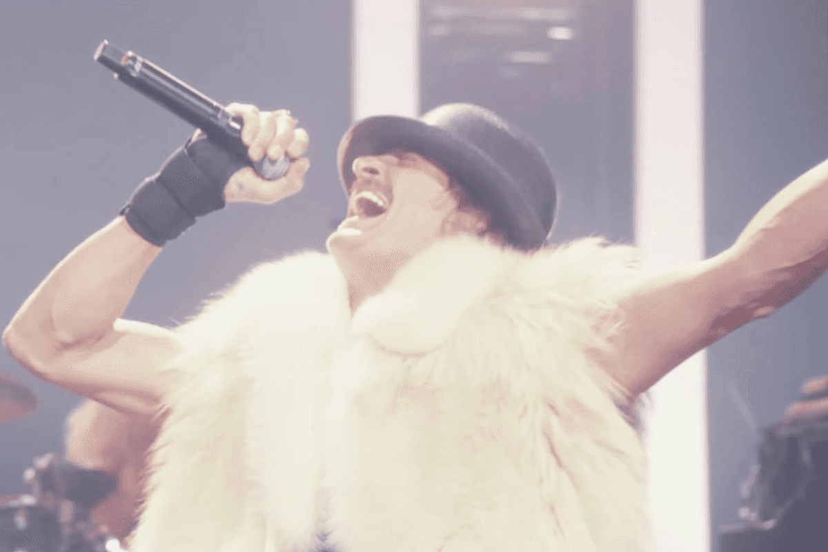 Kid Rock in a fluffy white fur jacket and black bowler hat, singing on stage.