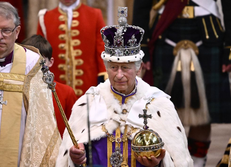 King Charles III, a white man with short grey hair and wearing royal clothing, departs the Coronation service at Westminster Abbey. He holds the orb and sceptre.