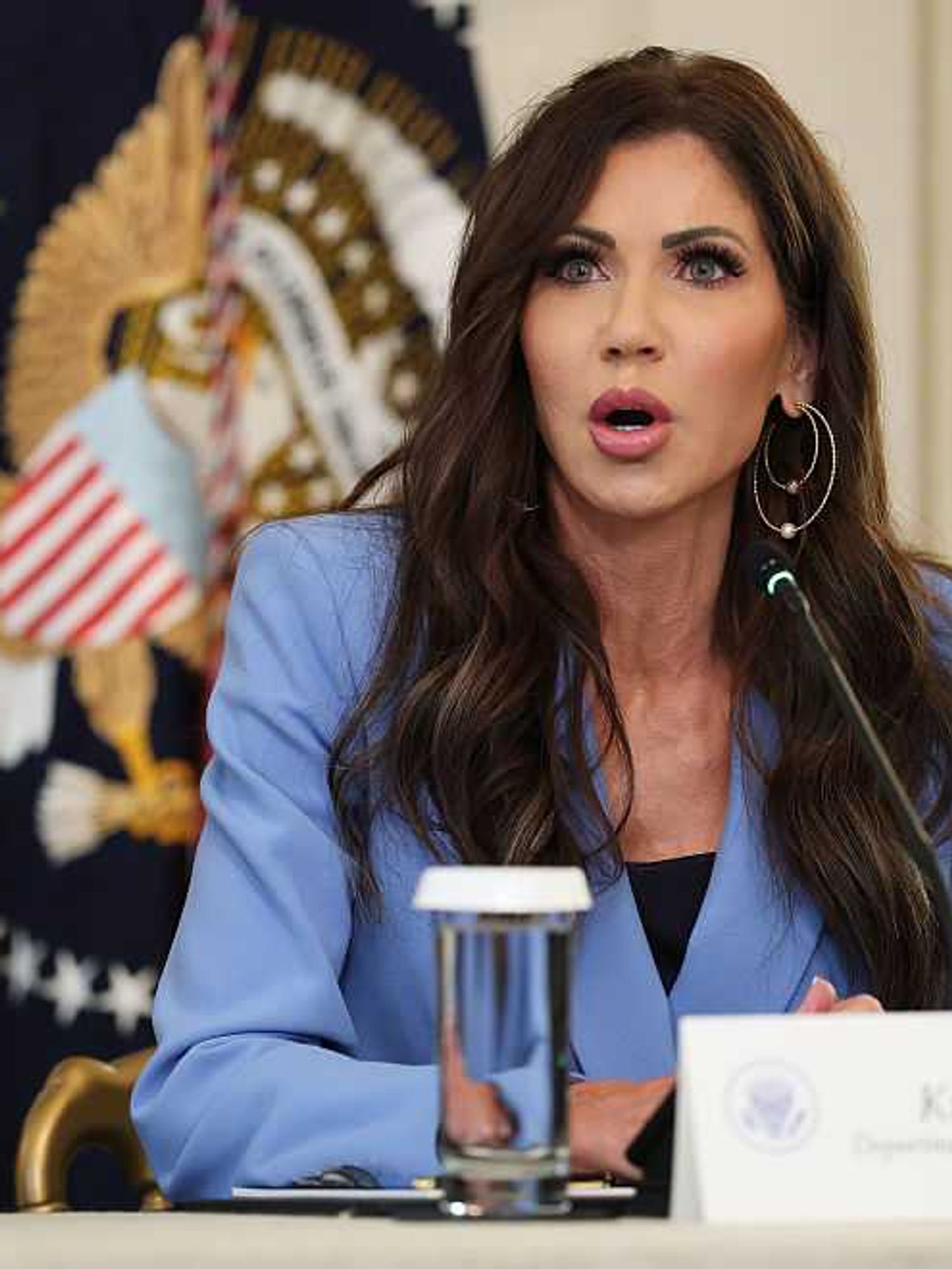 Kristi Noem sits behind a white desk wearing a light blue suit jacket.