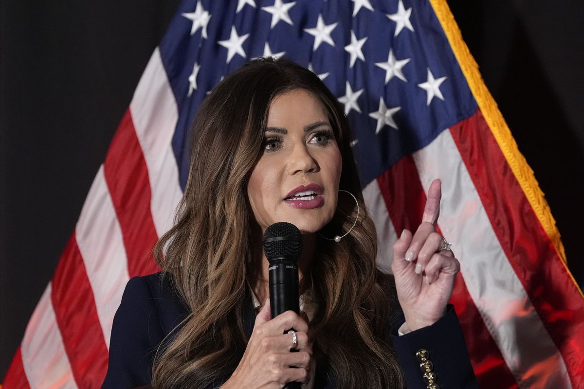Kristi Noem speaking into a microphone while standing in front of a US flag.