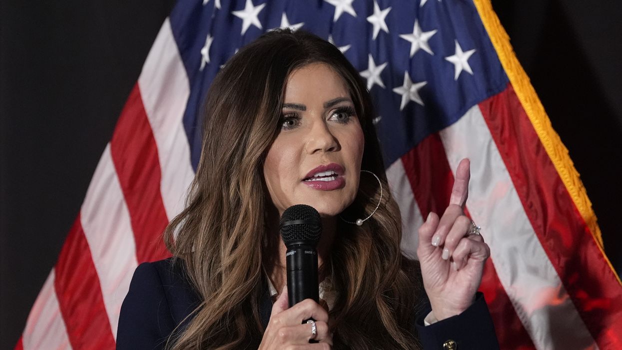 Kristi Noem speaking into a microphone while standing in front of a US flag.