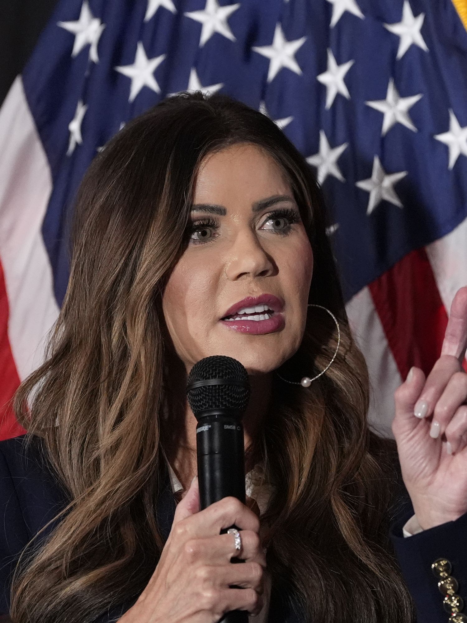 Kristi Noem speaking into a microphone while standing in front of a US flag.