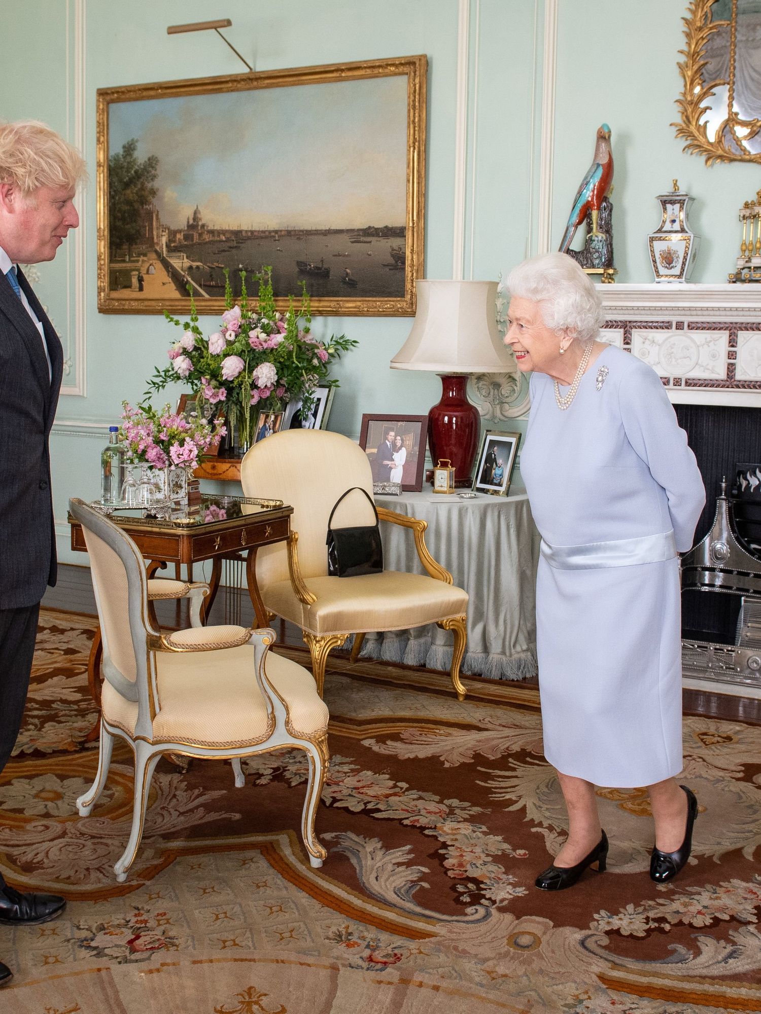 Left, Boris Johnson, a white man with scruffy blonde hair and a black suit, stands opposite the Queen, right. She's a white woman with grey hair and a lavender dress. They are in Buckingham Palace.
