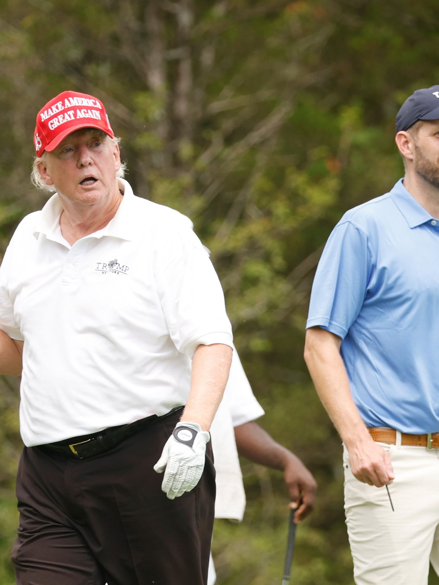 Left, Donald Trump, a white man with short blonde hair, a red MAGA cap and a white polo shirt, holds a golf club with his mouth open. Right, Eric Trump wears a blue polo shirt and blue cap.