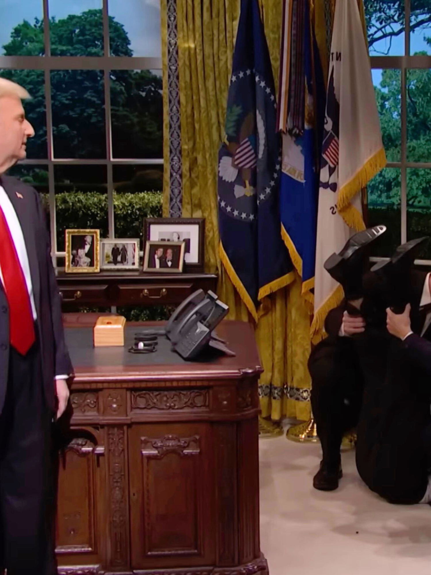 Left, James Austin Johnson on SNL as Donald Trump, turning to his left to look behind him at several men tending to a man on the floor on the right, lifting his feet in the air.