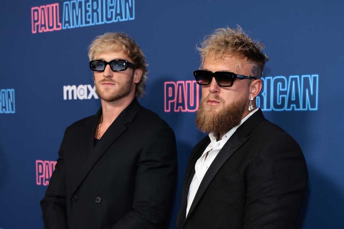 Left, Logan Paul, and right, Jake Paul, wearing black suits and sunglasses in front of a blue posterboard.