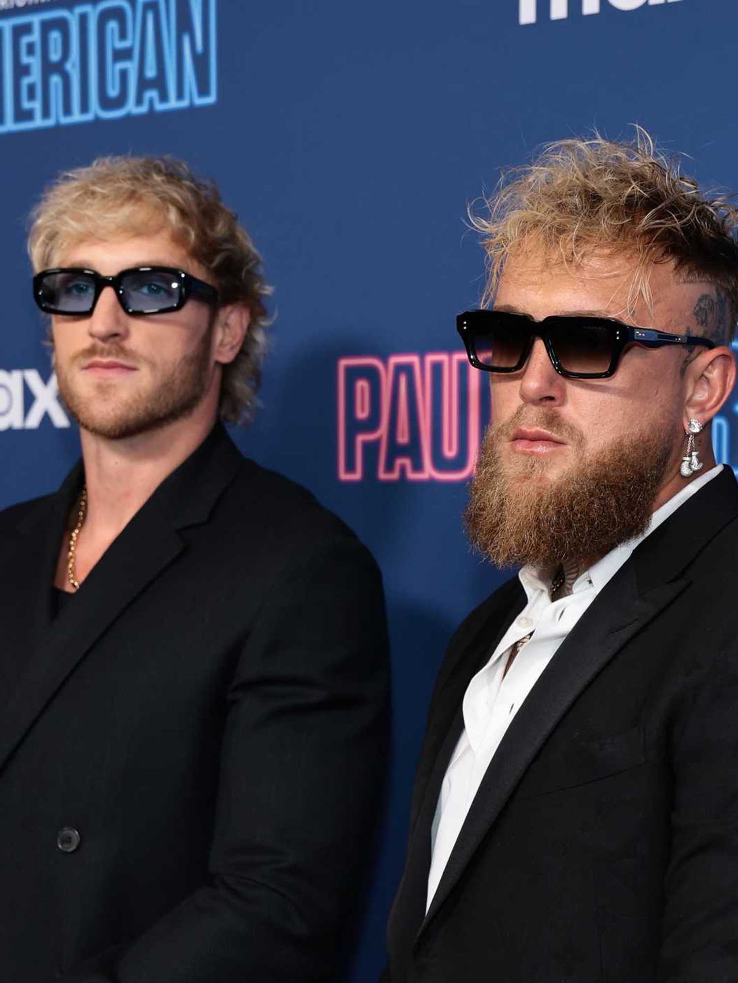 Left, Logan Paul, and right, Jake Paul, wearing black suits and sunglasses in front of a blue posterboard.