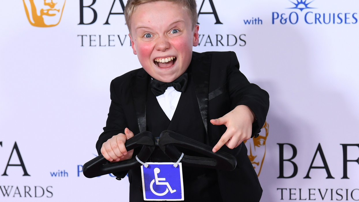 Lenny Rush, a white teenager of short stature with short brown hair and a black suit, smiles while on a segway in front of a white Bafta posterboard.