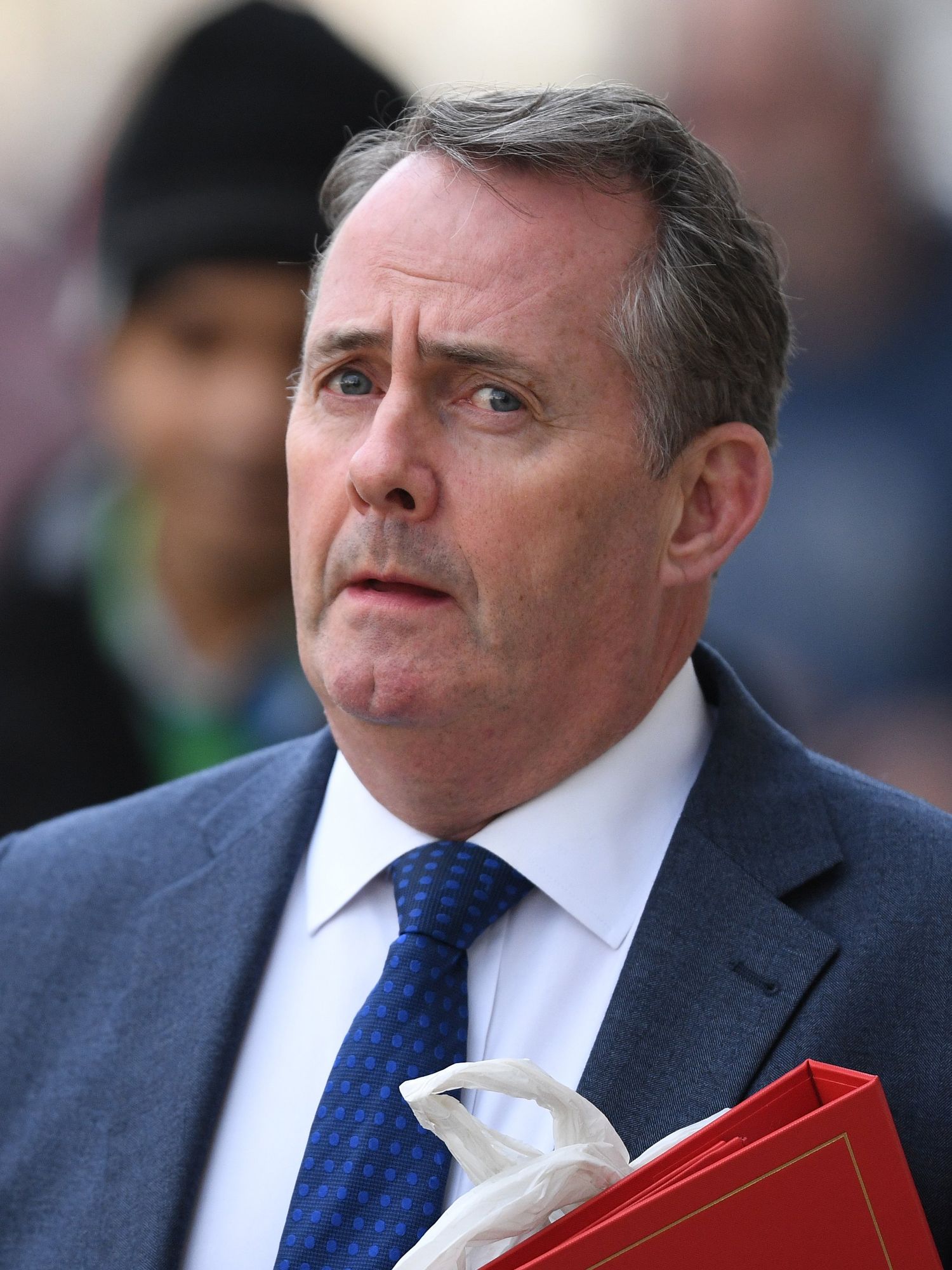 Liam Fox, holding a red folder and wearing a blue suit, looking at the camera with a neutral expression on his face.