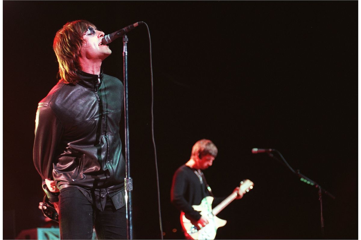Liam Gallagher leans into the microphone as he performs on stage with Oasis. He is wearing a leather jacket and sunglasses.