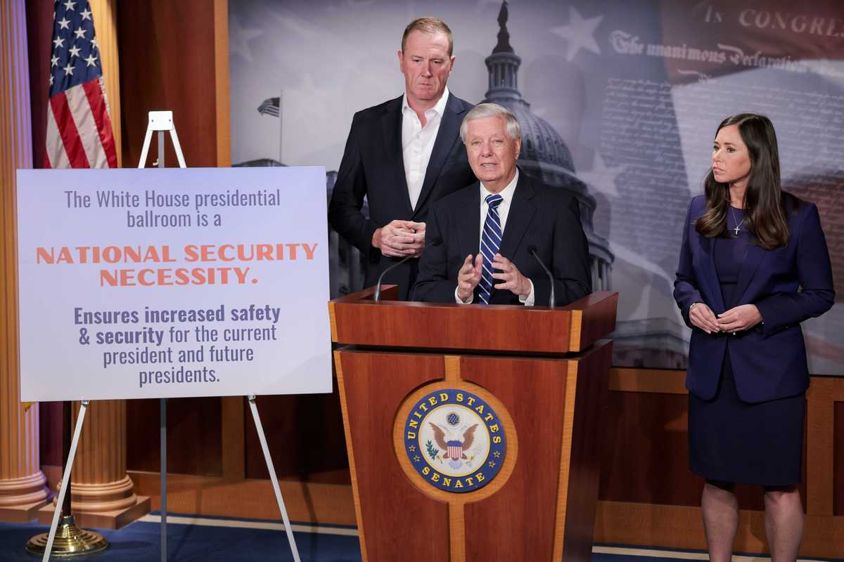 Lindsey Graham, a white man with short grey hair, speaks behind a wooden lectern. A white man and white woman stand behind him. A canvas on his right displays the text: 'The White House presidential ballroom is a national security necessity. Ensures increased safety & security for the current president and future presidents.'