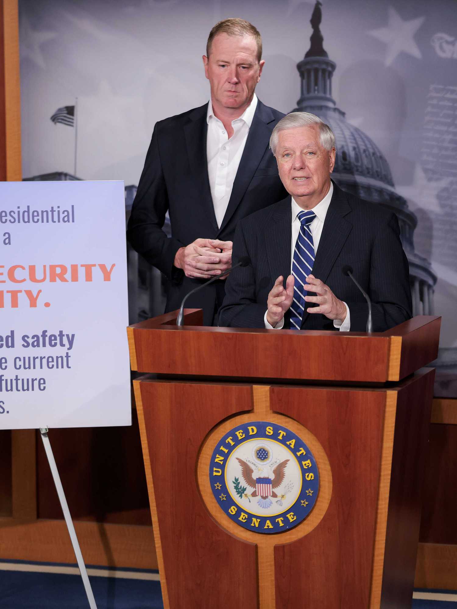 Lindsey Graham, a white man with short grey hair, speaks behind a wooden lectern. A white man and white woman stand behind him. A canvas on his right displays the text: 'The White House presidential ballroom is a national security necessity. Ensures increased safety & security for the current president and future presidents.'