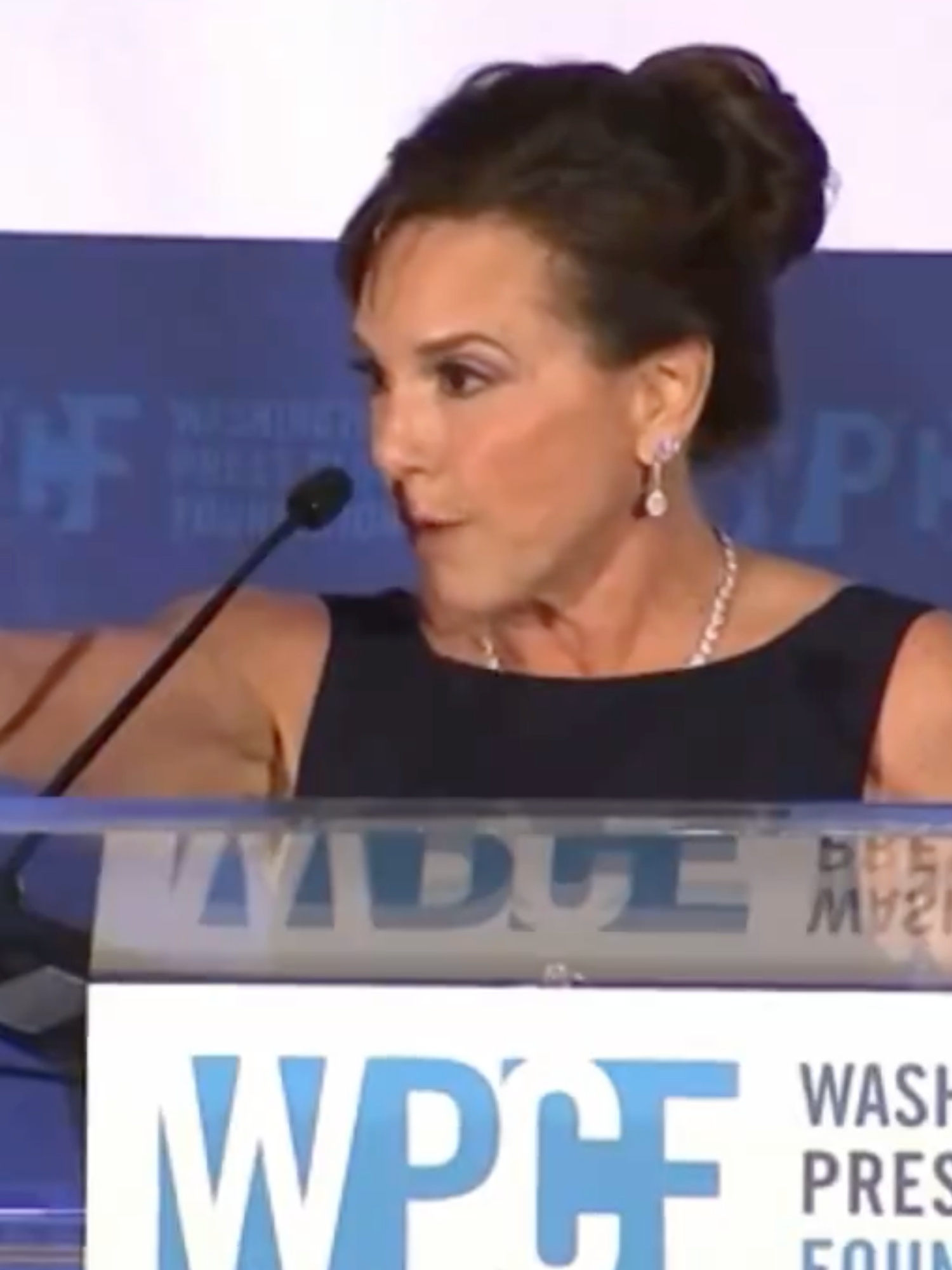 Lisa McClain, a white woman with her black hair tied back, stands behind a lectern and has her right hand out pointing to someone in the crowd in front of her.