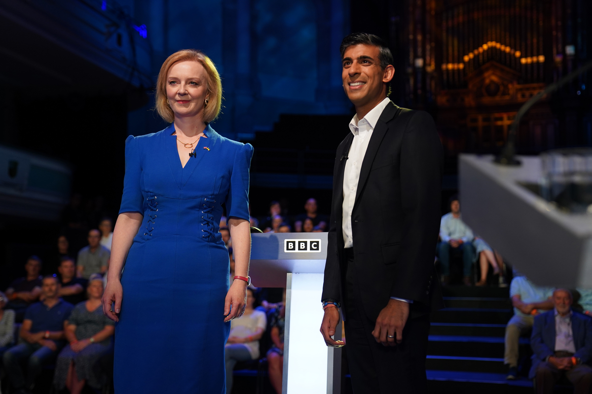 Liz Truss, a white woman with short blonde hair and a blue dress, and Rishi Sunak, a brown man in a black suit, both smile at the camera as they stand on stage with an audience seated behind them. it's a TV debate.
