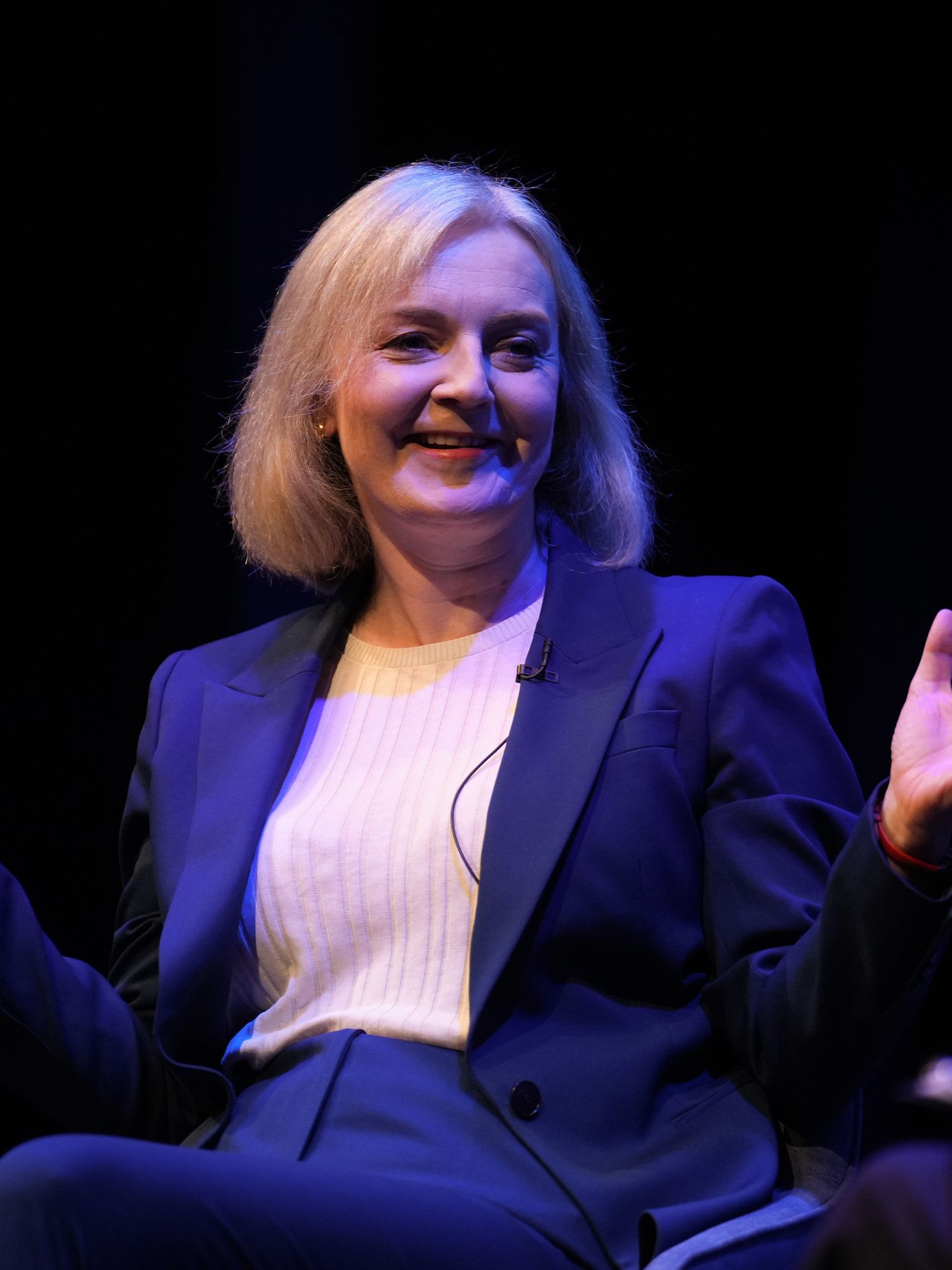 Liz Truss, a white woman with short blonde hair and a blue suit, sits on a chair and raises both hands upwards, as if making a shrugging gesture. She's smiling.