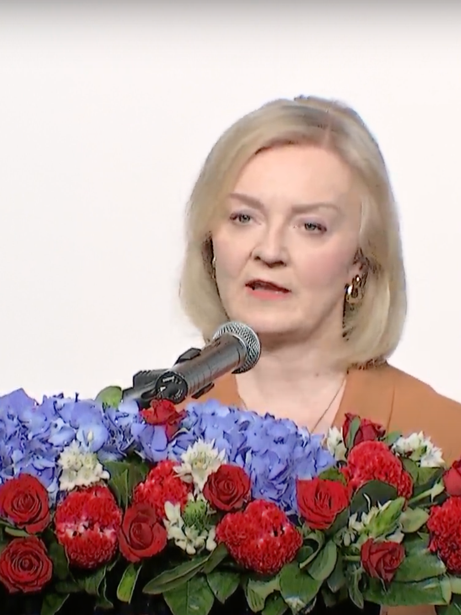 Liz Truss, a white woman with short blonde hair and an orange dress, speaks behind a floral lectern.