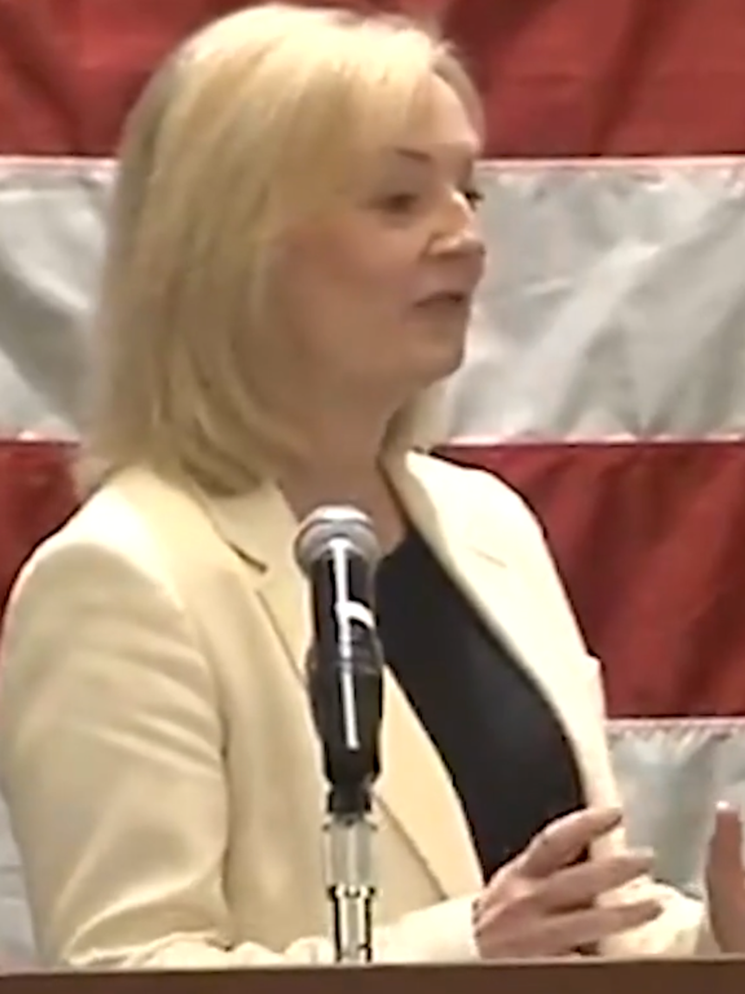 Liz Truss, in a white suit, delivering a speech in front of an American flag.