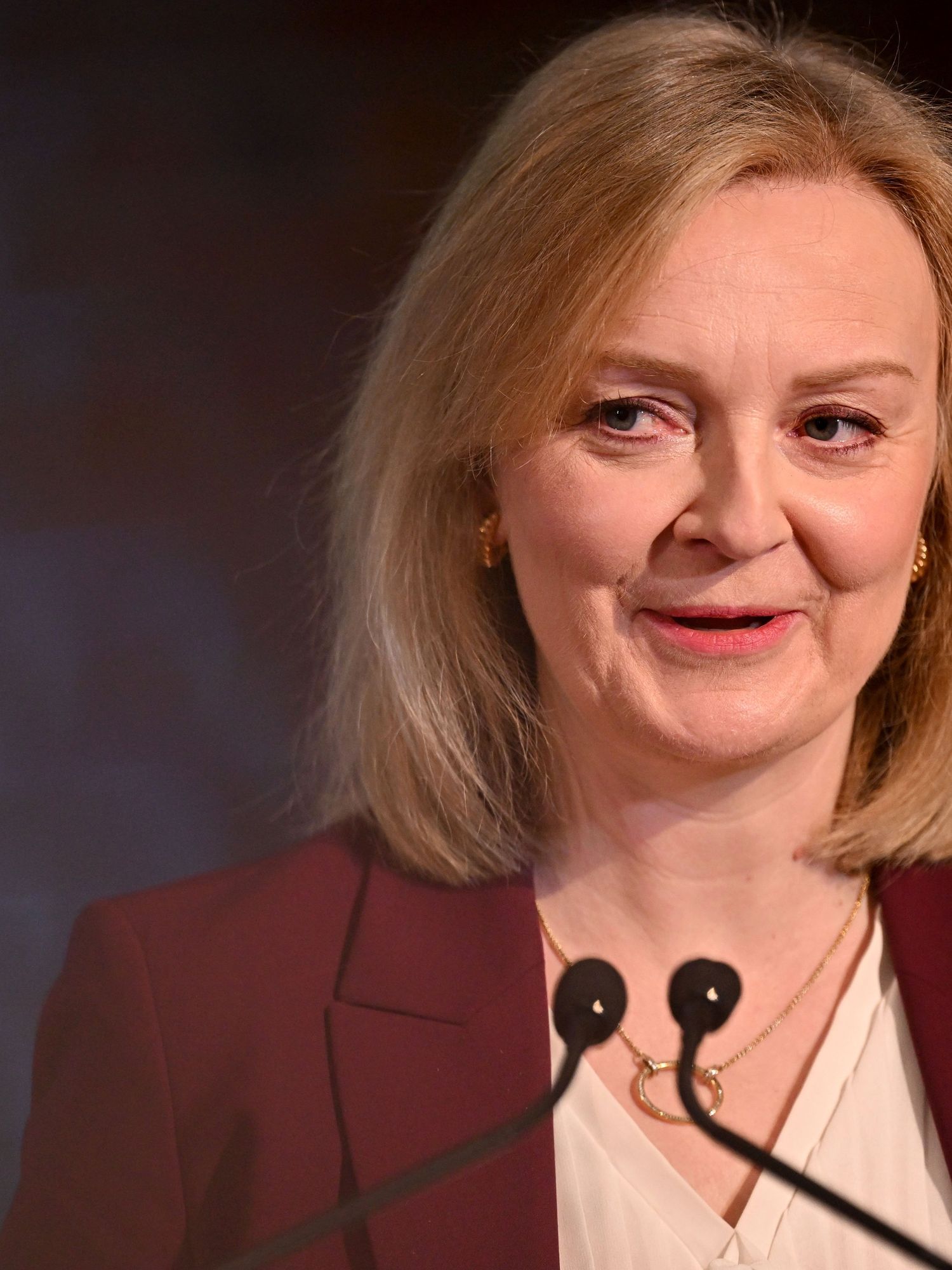 Liz Truss wearing a maroon suit jacket and speaking in front of a lectern.