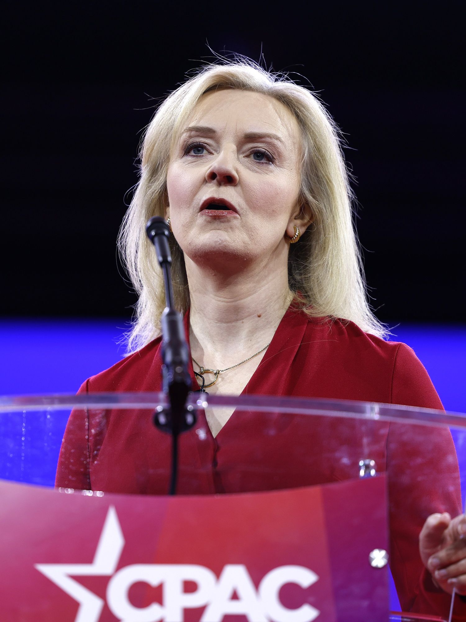 Liz Truss wearing a red dress and speaking in front of a clear lectern with a red CPAC logo on it.