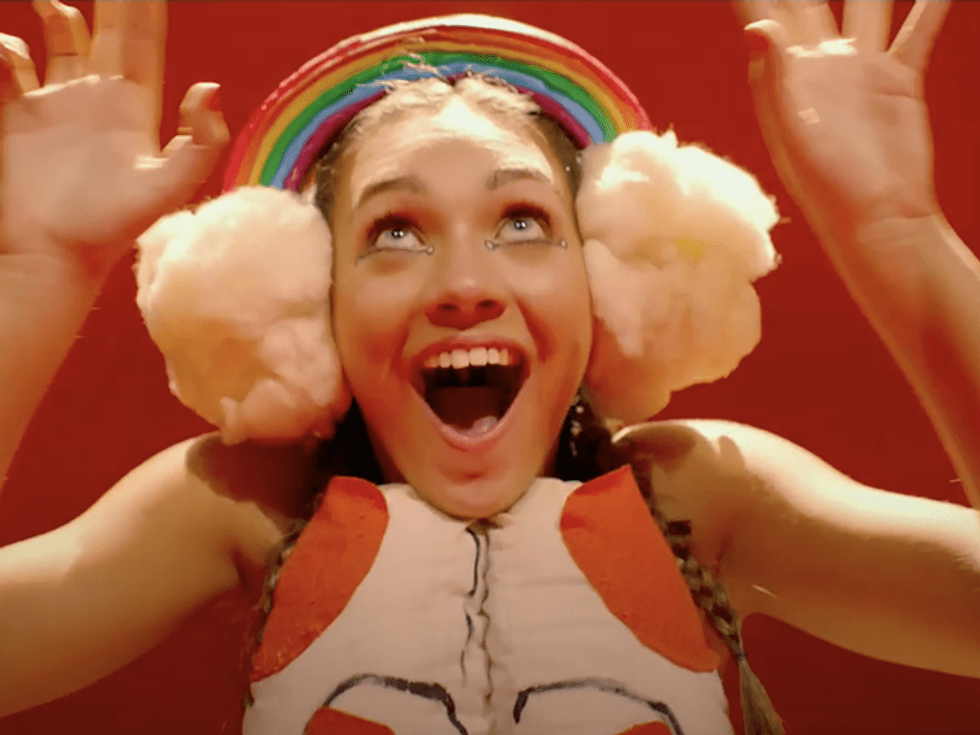 Maddie Ziegler in the film Music. Her mouth and eyes are opened wide, and she is smiling. On her head is a rainbow, with two clouds either side which look like earmuffs.